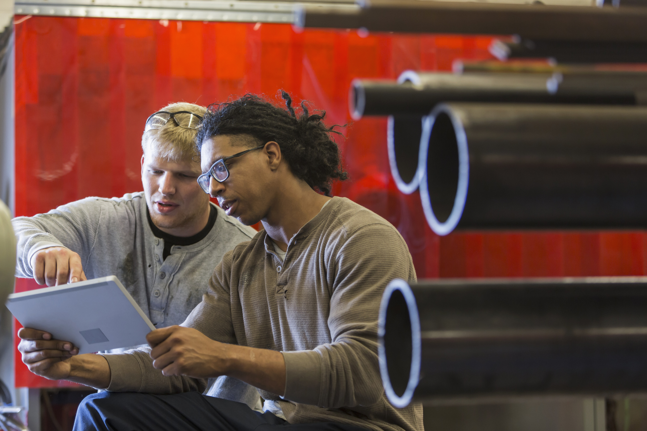Two men sit in an industrial setting, examining a digital tablet together, with large metal pipes visible in the foreground.