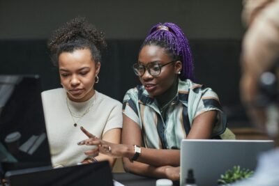Two women sit side by side at a desk, looking at a laptop screen while one gestures and discusses something with the other.