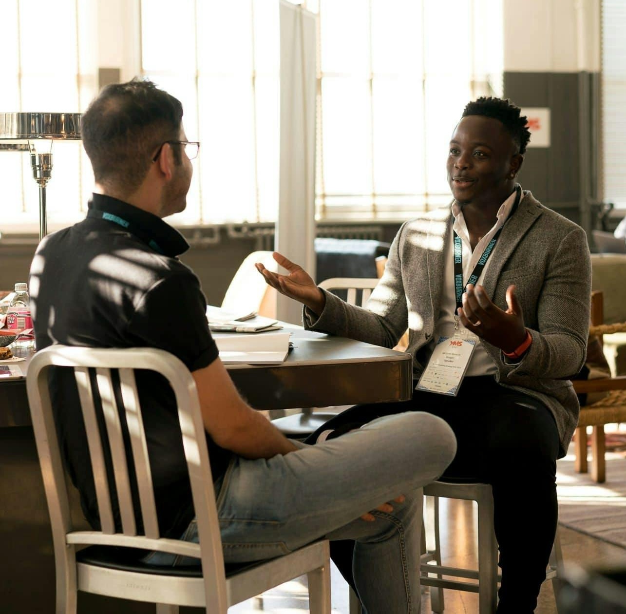 Two men sit at a table in conversation, both wearing lanyards and name tags, in a well-lit room with large windows in the background.