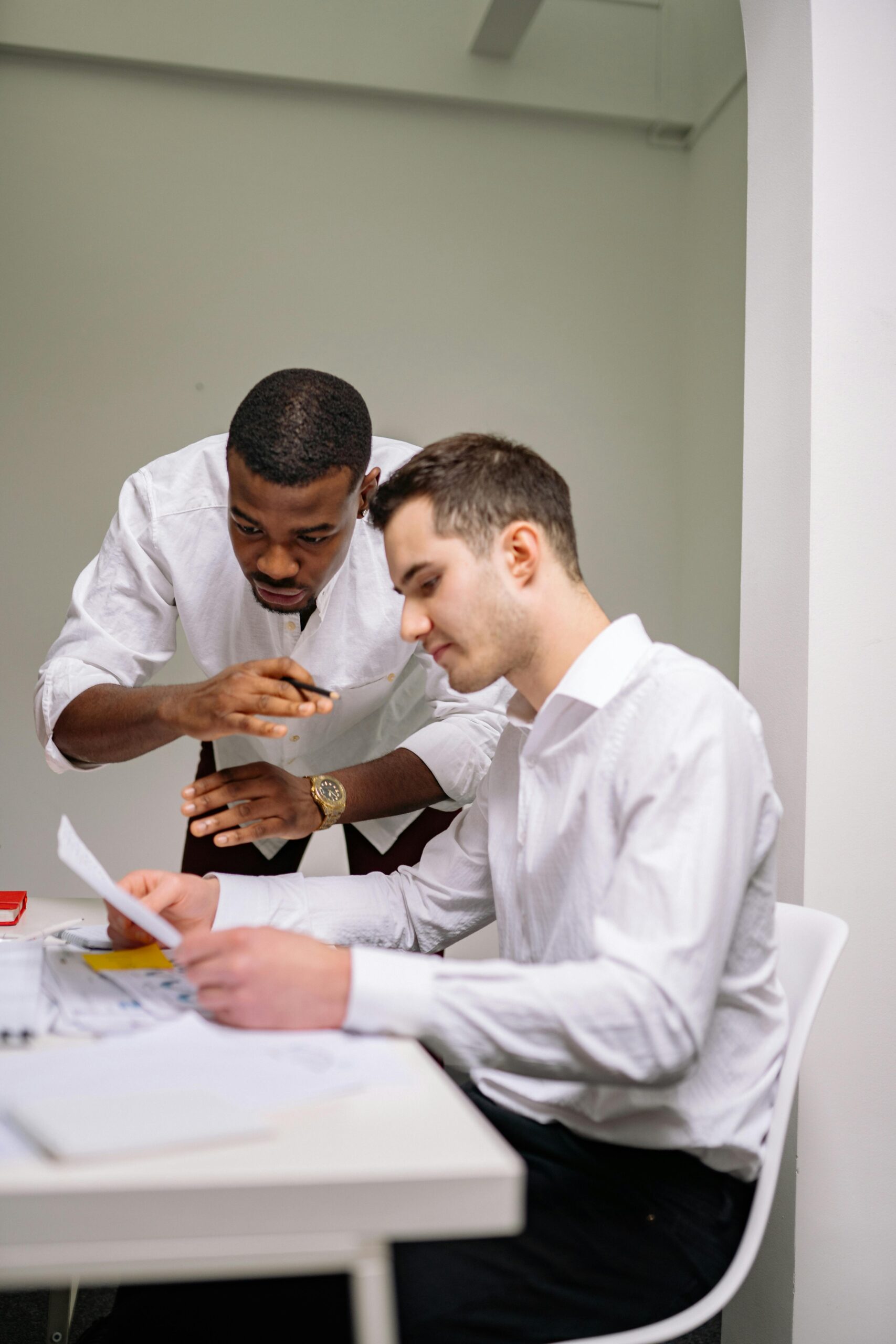 Two men in white shirts review documents together at a desk in a modern office setting. One is standing and pointing, while the other sits and reads the papers.