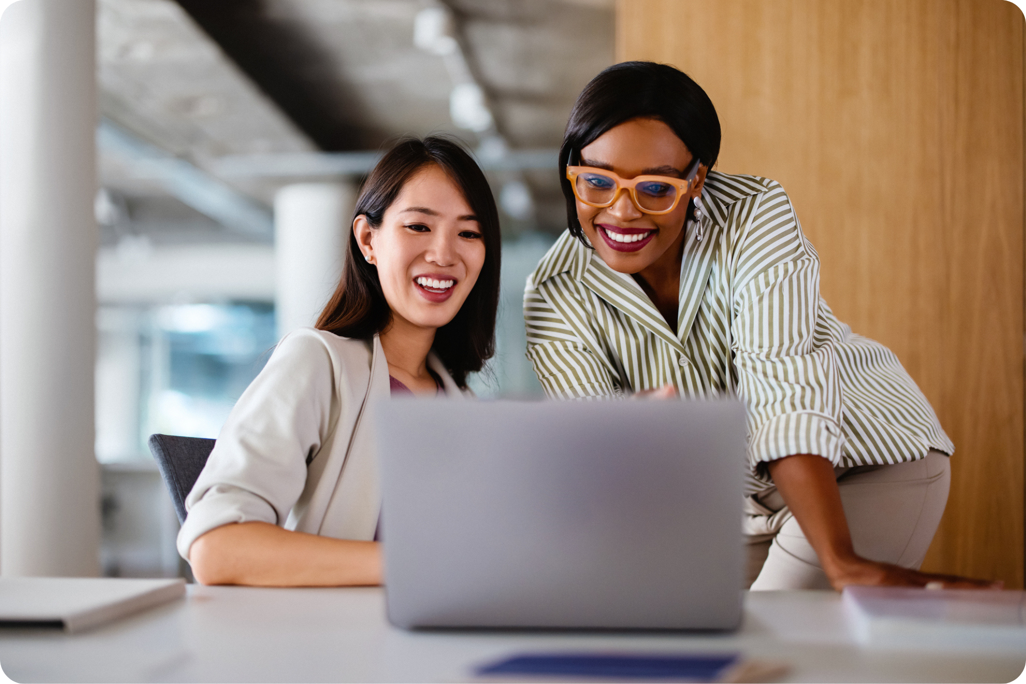 Two women in business attire smiling and looking at a laptop screen in a modern office setting.