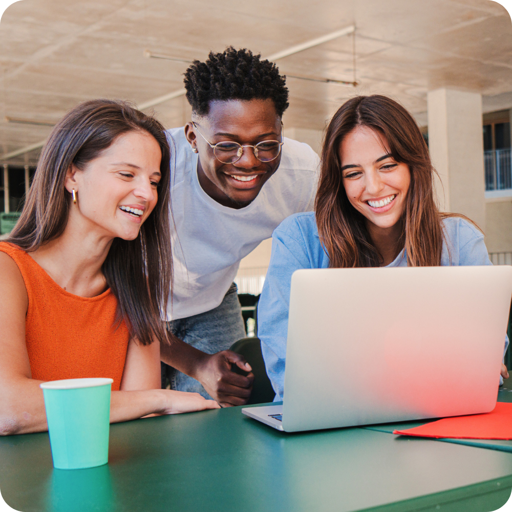 Three young adults smile while looking at a laptop on a green table; a paper cup and red folder sit nearby in an indoor setting.