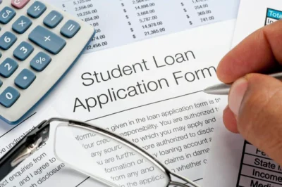 A close-up of a hand filling out a student loan application form, with a calculator, eyeglasses, and financial documents nearby.