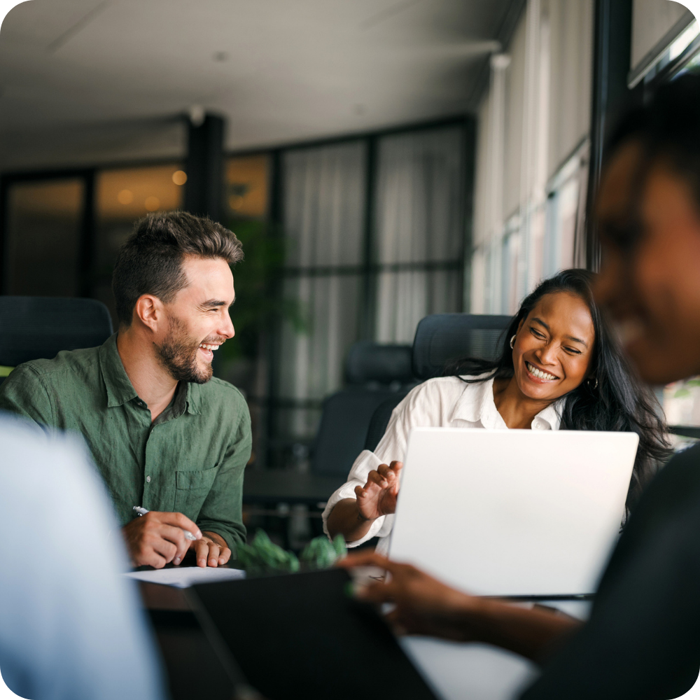 Two people sit at a table in an office, smiling and talking while working on a laptop, with others blurred in the foreground.