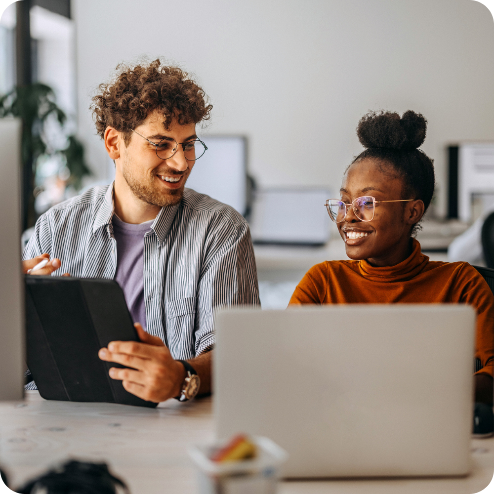 Two colleagues sit at a desk with laptops, smiling and discussing something on a tablet in a modern office setting.