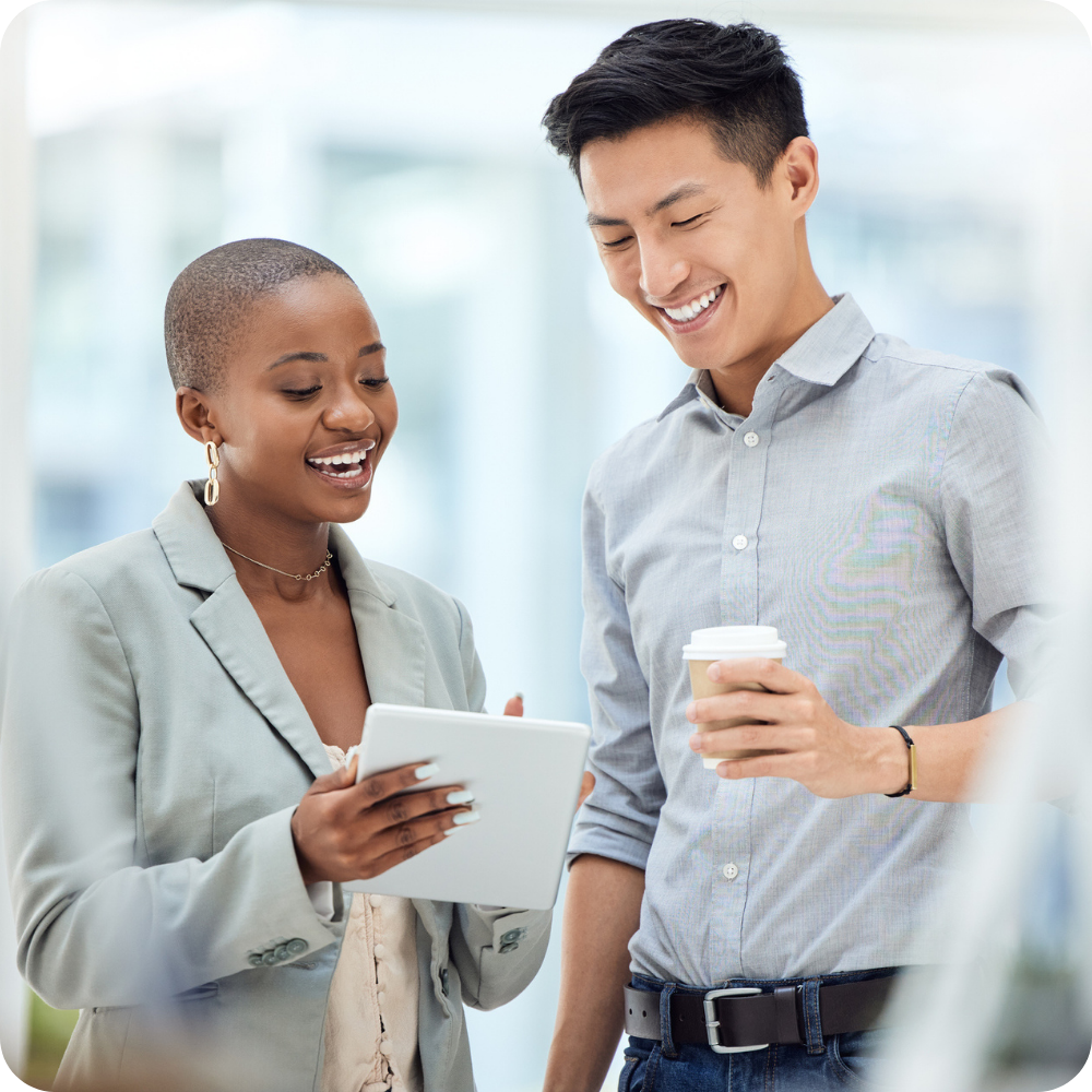 Two people standing indoors, one holding a tablet and showing it to the other, who is smiling and holding a disposable coffee cup.