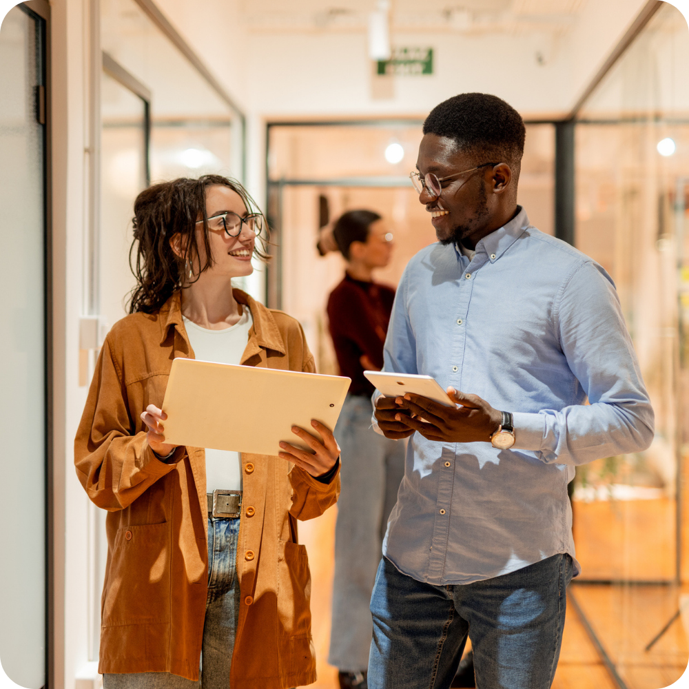 Two colleagues walk down an office hallway, smiling and holding digital tablets, while another person stands in the background.