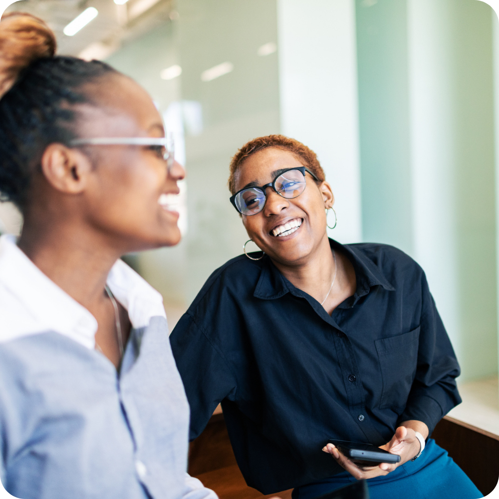 Two people wearing glasses are smiling and talking indoors. One person is holding a mobile phone. The background is bright and modern.
