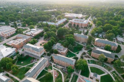 Aerial view of a college campus, brick academic buildings, green lawns, walking paths, and surrounding trees.