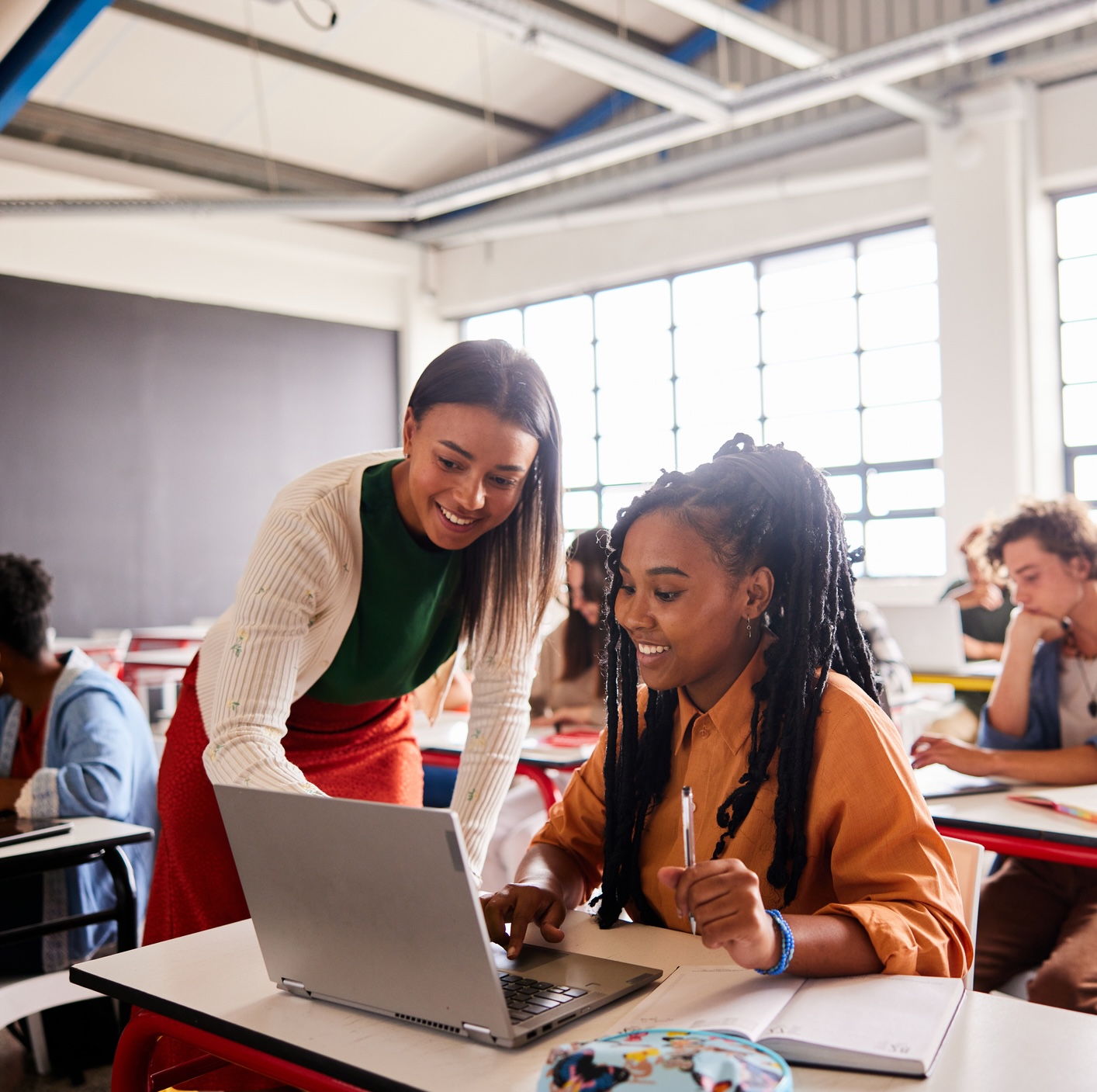 A teacher helps a student with a laptop in a classroom, while other students work at their desks in the background.