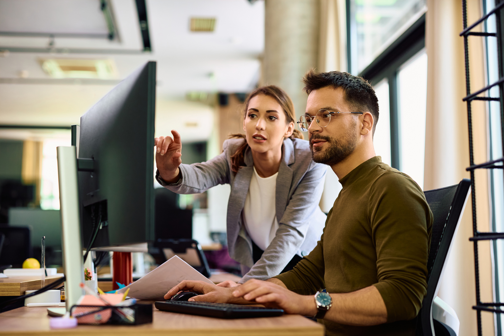 Two colleagues working at a computer desk; one is seated typing while the other stands beside him, pointing at the monitor and discussing something in an office setting.