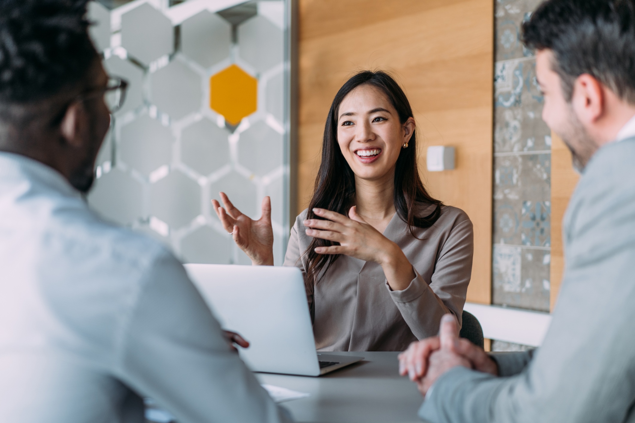 Three people in business attire sit at a table in an office, having a discussion. One woman is talking and gesturing with her hands in front of an open laptop.