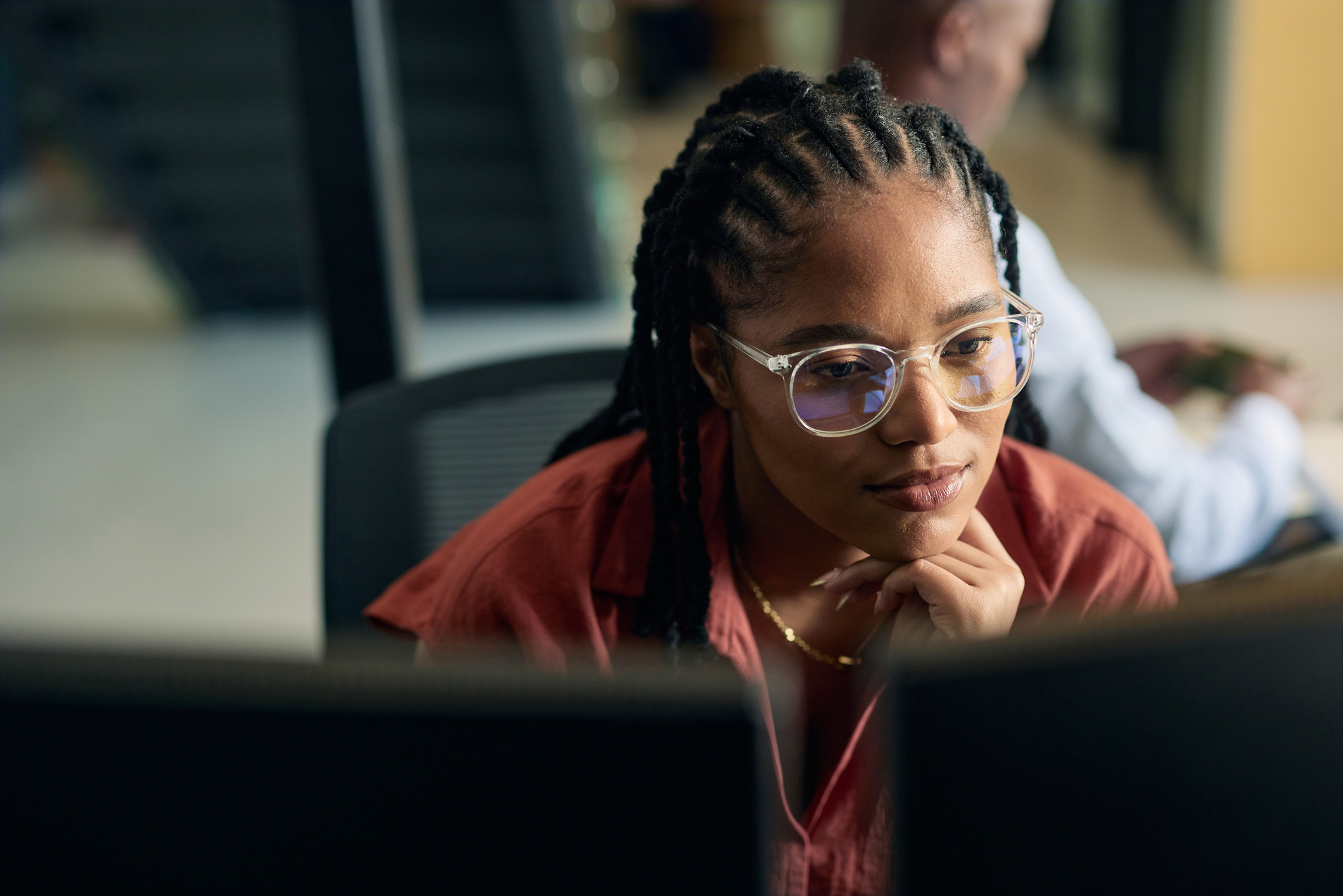 A woman with braided hair and glasses looks intently at a computer screen in an office setting.