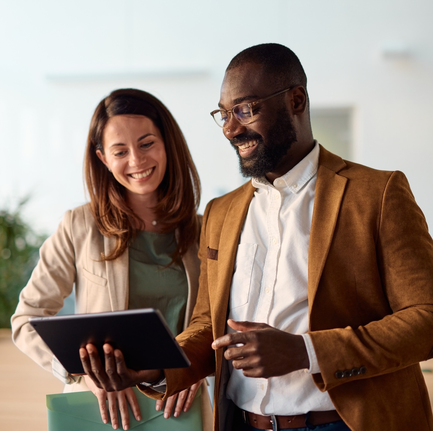 Two professionally dressed people stand indoors, smiling and looking at a tablet that one of them is holding.
