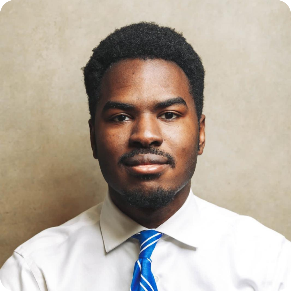 A man with short black hair and a trimmed beard wears a white shirt and blue striped tie, posing against a plain beige background.