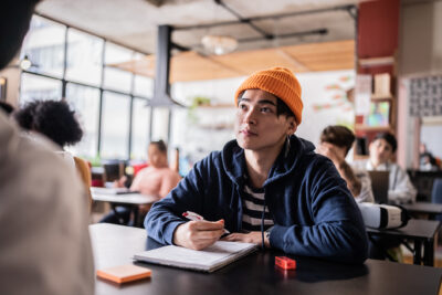 A young man in a striped shirt and orange beanie listens attentively while taking notes in a busy classroom setting.