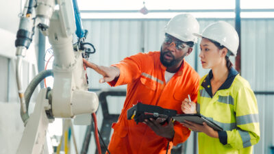 Two engineers in safety gear examine and discuss an industrial robotic arm, holding tablets and clipboards in a factory setting.