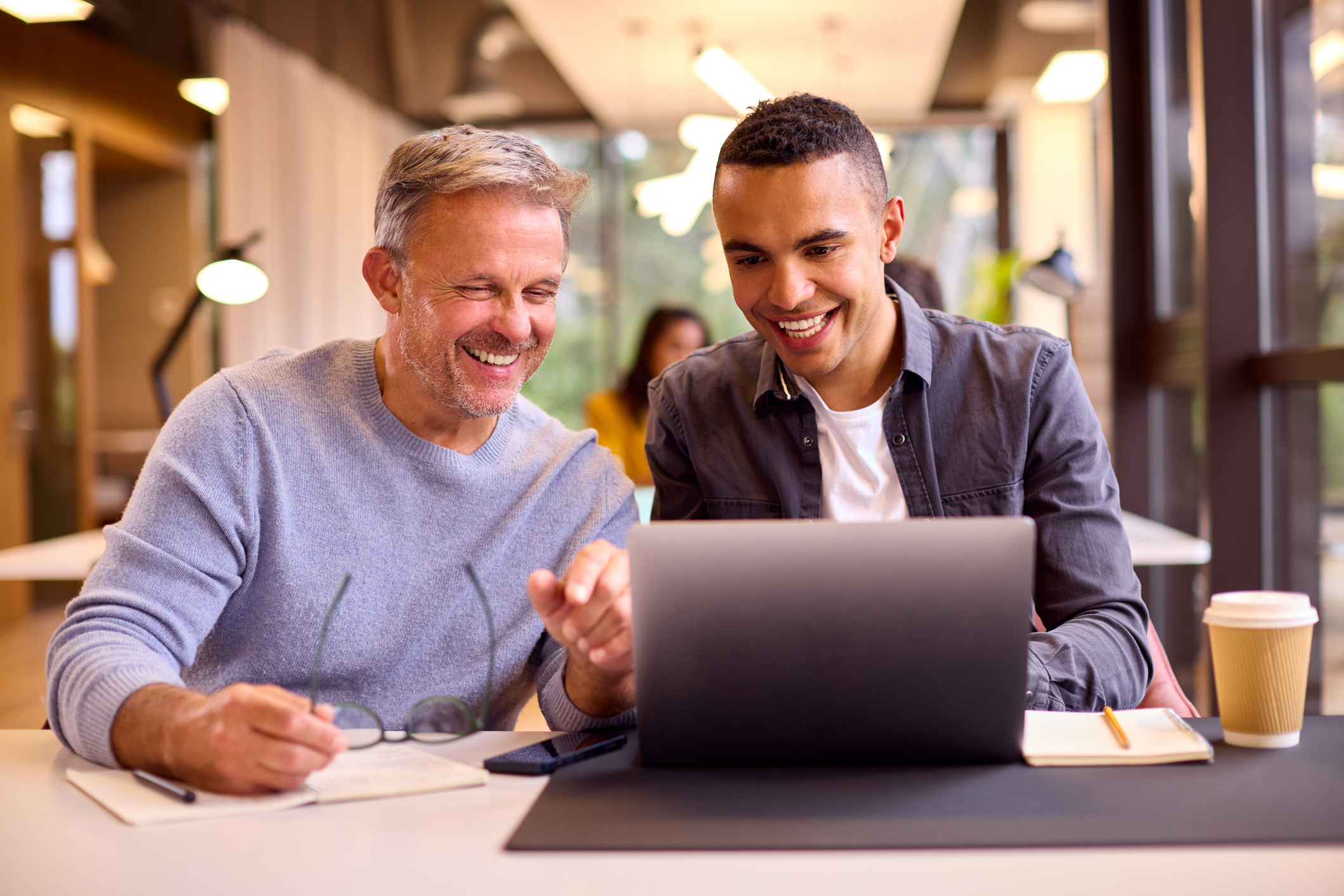 Two men sit at a table, smiling and looking at a laptop screen together, with notepads and a coffee cup on the table.
