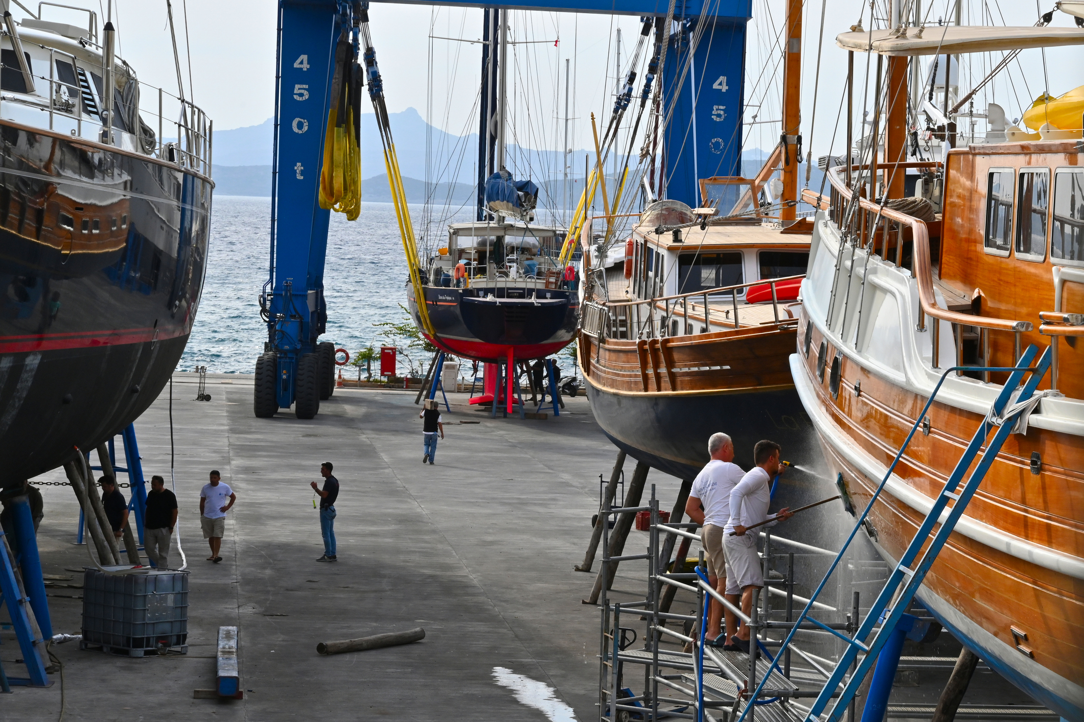 Several boats are docked onshore at a marina, with people performing maintenance near a large blue boat lift crane by the sea.