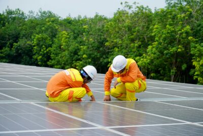 Two workers wearing helmets and safety uniforms inspect solar panels outdoors, surrounded by green trees.