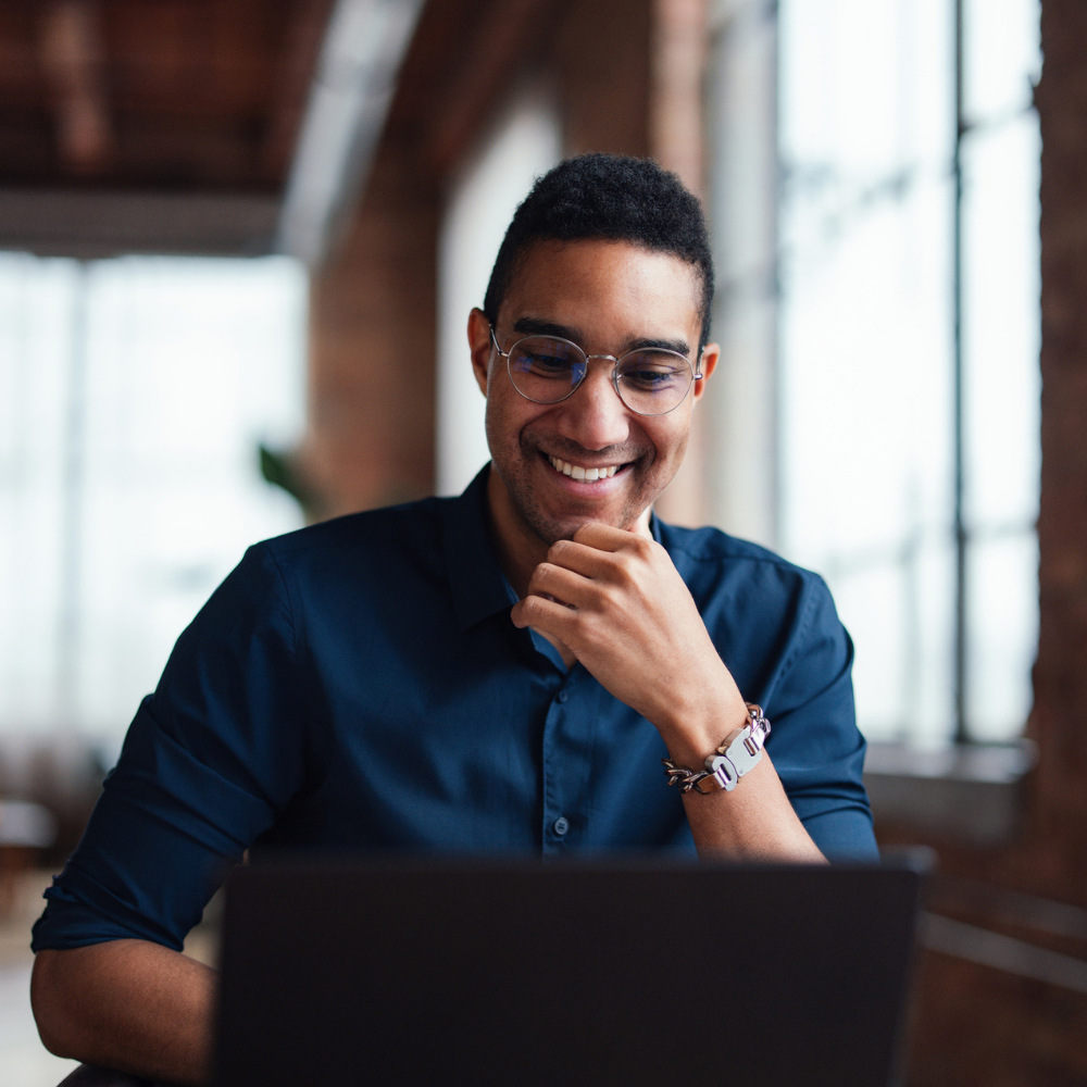 A man wearing glasses and a dark shirt sits at a desk, smiling while looking at a laptop in a bright, modern office space.