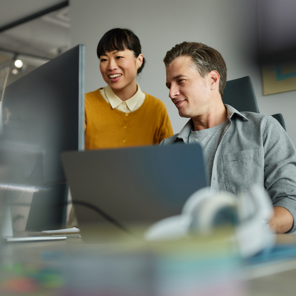 Two people working together at a desk, looking at a computer monitor. One person is seated while the other stands beside them, both focused on the screen.