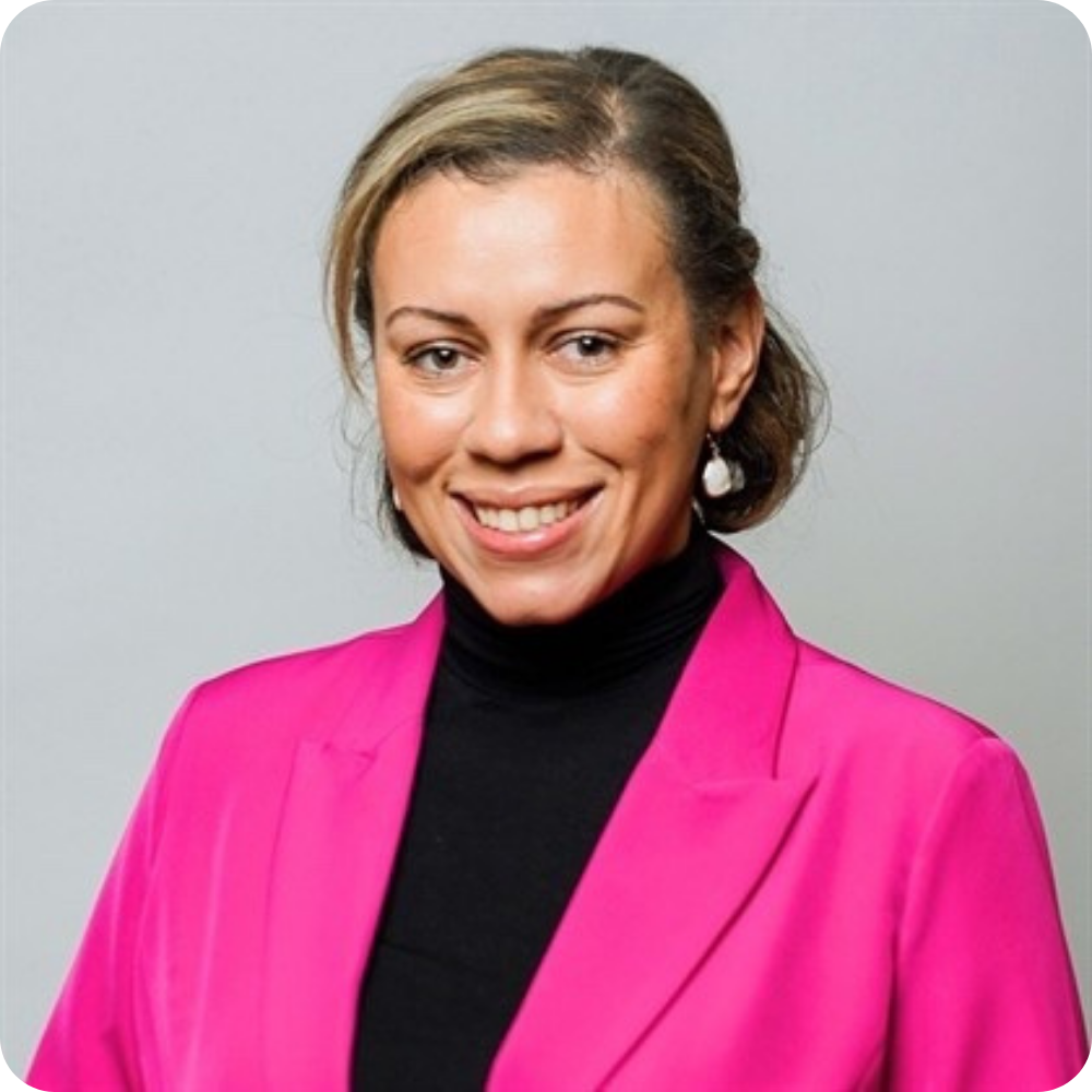 A woman with light brown hair in an updo wears a bright pink blazer over a black top, smiling at the camera against a plain gray background.