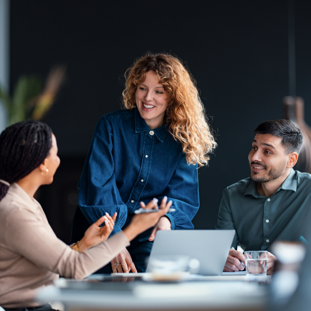 Three people have a discussion around a table with a laptop, notepads, and a glass of water in an office setting.