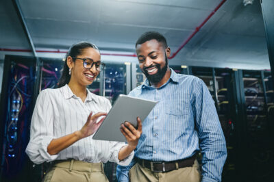 Two professionals standing in a server room look at a tablet together, with server racks and network cables visible in the background.