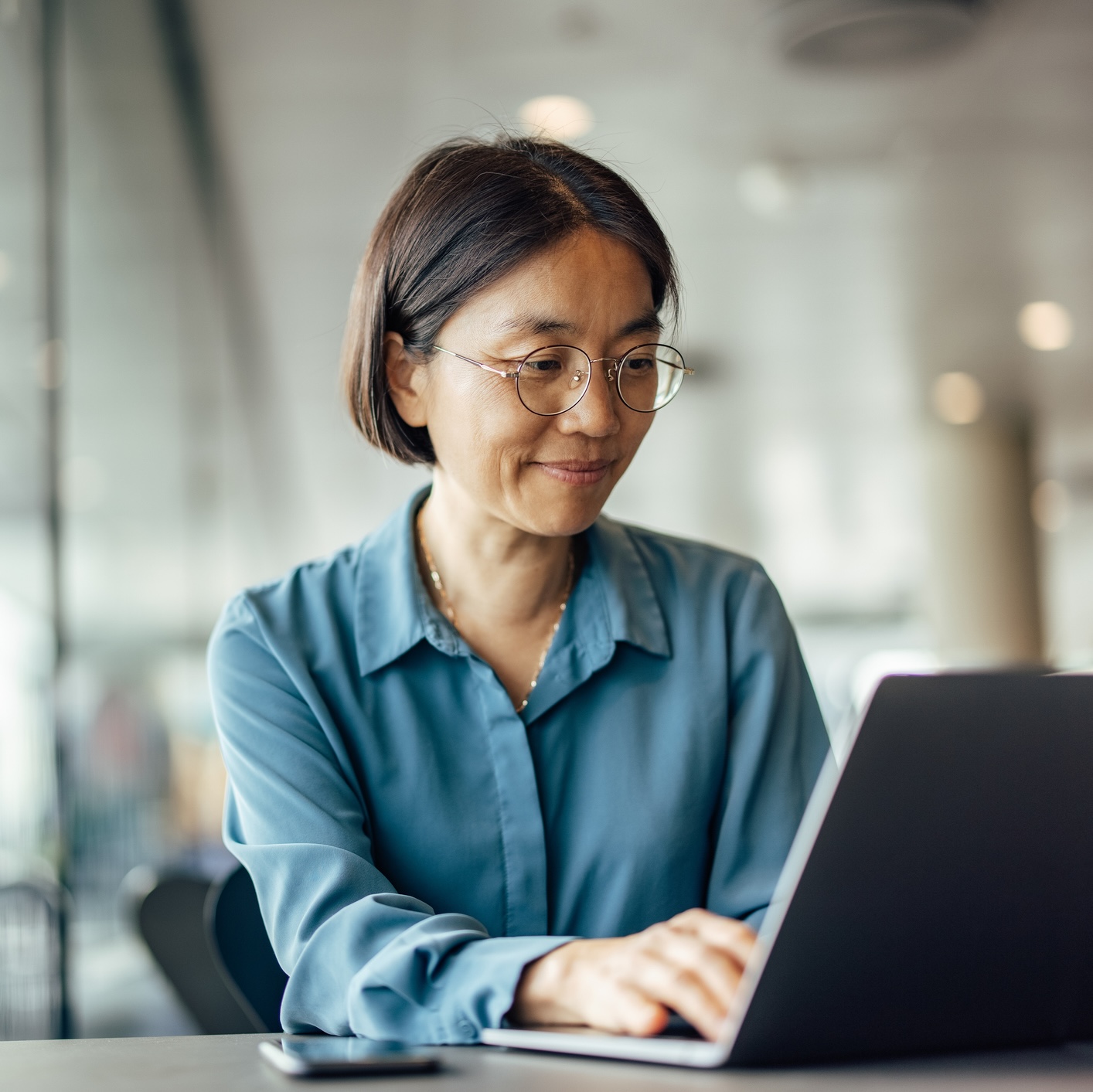 Woman in glasses and a blue blouse working on a laptop at a table in a bright indoor setting, with a smartphone beside her.