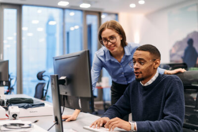 Two colleagues in an office look at a computer screen together; one person is seated and typing while the other stands and observes.