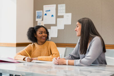 Two women sit at a table in a meeting room, facing each other and talking. One has a notebook open. Papers and charts are on the wall in the background.
