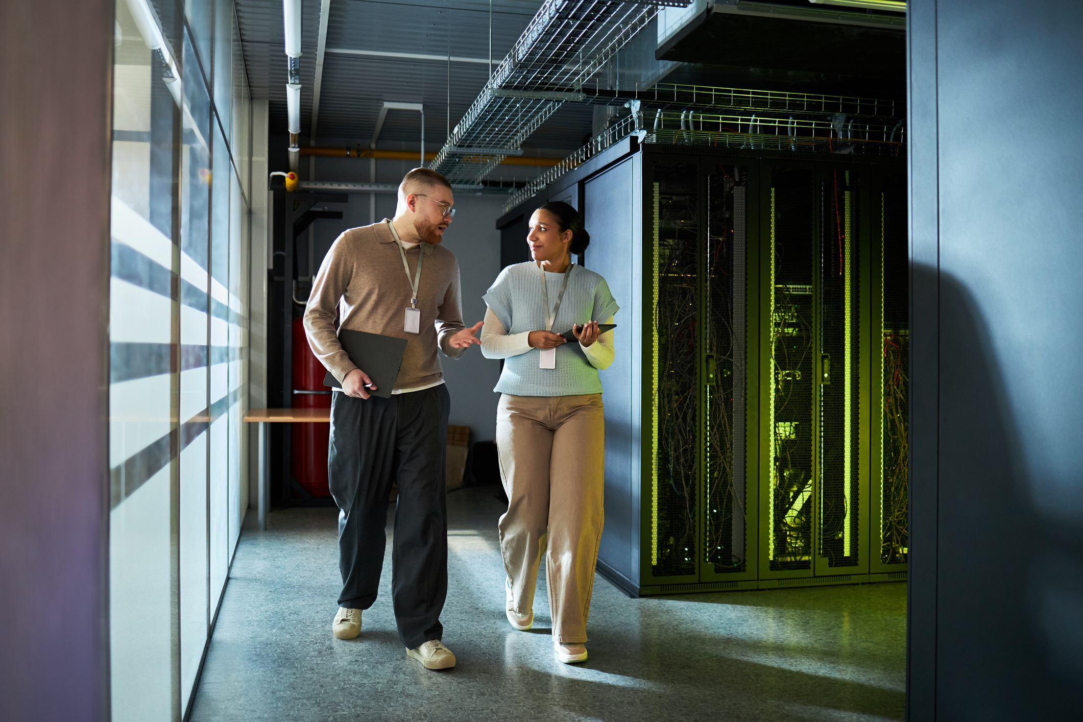 Two IT professionals walk and talk in a data center hallway, carrying a laptop and tablet, with server racks visible in the background.