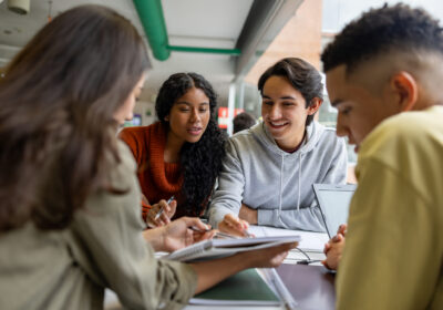 Four young adults sit at a table with books and a laptop, collaborating and discussing a notebook in a bright indoor setting.