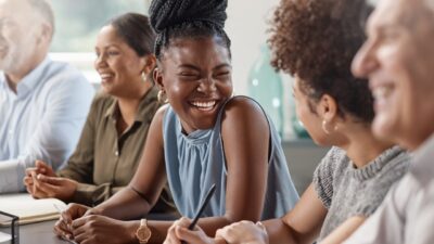 Four people sit at a table, smiling and talking together. The focus is on a woman in a blue sleeveless top, laughing with others in a well-lit, casual setting.