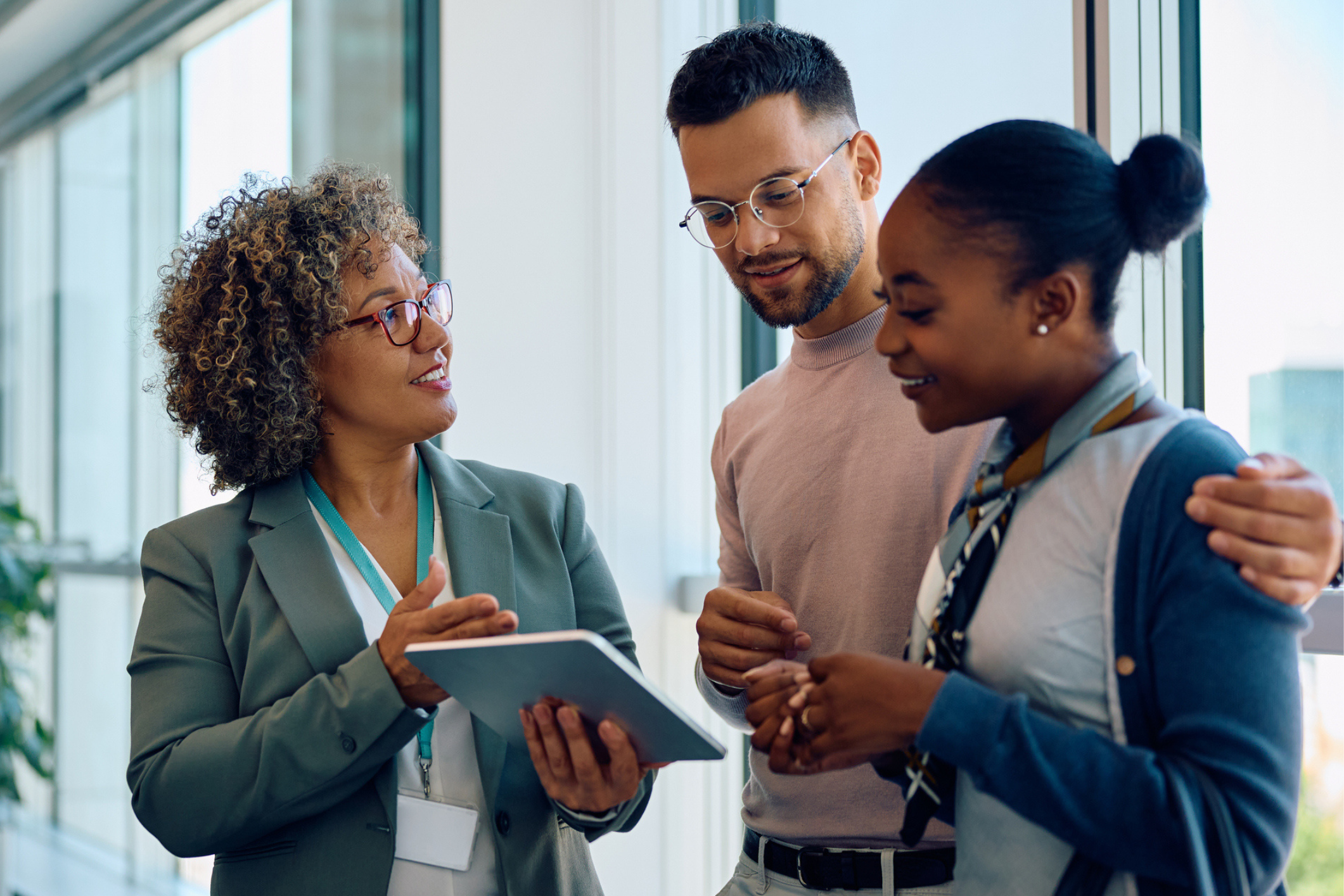 Three people stand by a window, looking at a digital tablet together. One person holds the tablet and speaks, while the other two listen and smile.