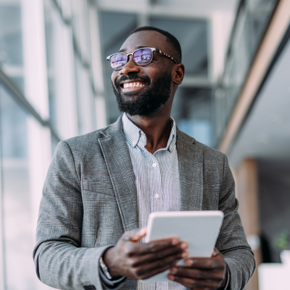 A man wearing glasses and a gray blazer holds a tablet and smiles while standing in a modern, glass-walled building.