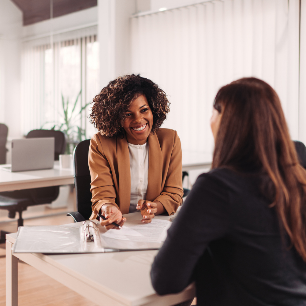Two women sit across from each other at a desk in an office, discussing documents laid out on the table.