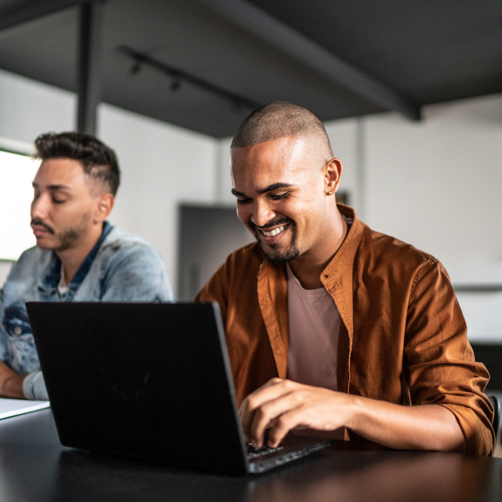 Two men sit at a table with laptops; one man smiles while typing, the other looks at his screen.