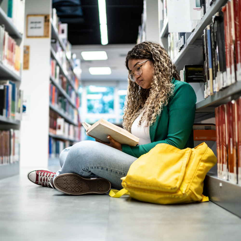 A woman sits on the floor between library shelves, reading a book. A yellow backpack rests beside her.