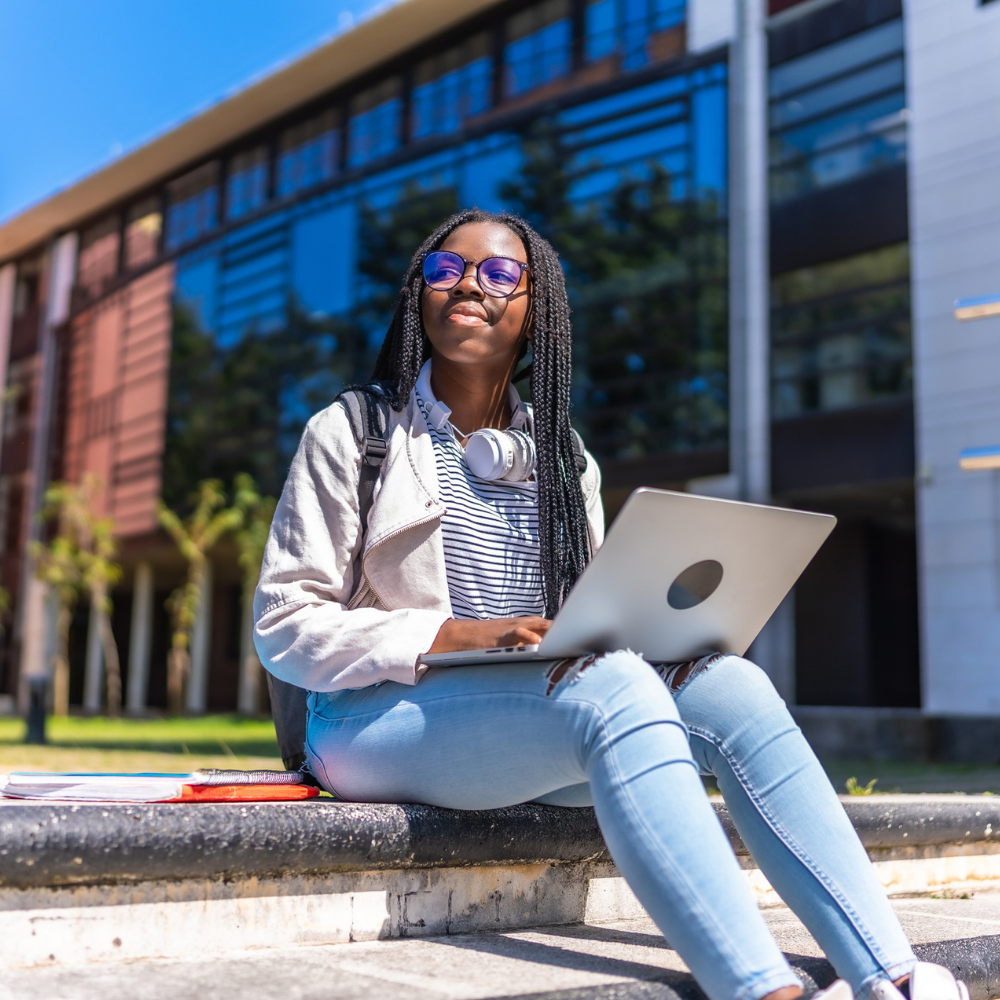 A young woman with braided hair and glasses sits outdoors on a curb, using a laptop. She has headphones around her neck and a backpack, and is in front of a modern building.