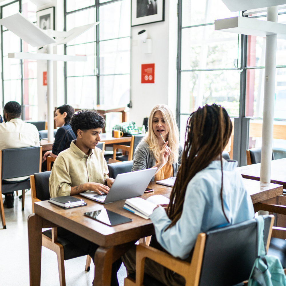 Three people sit at a table in a library, talking and using laptops and notebooks.