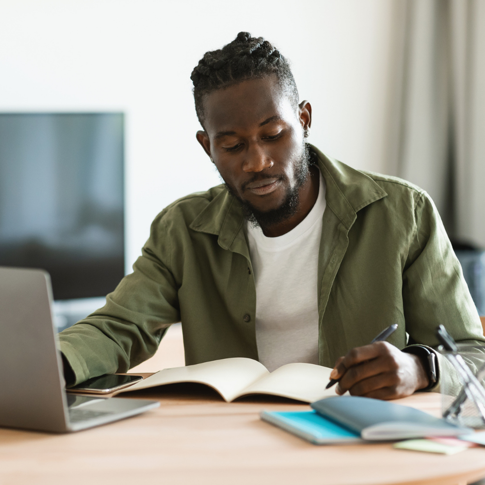 A man sits at a desk with a laptop, writing in a notebook. Other notebooks and a smartphone are on the table.