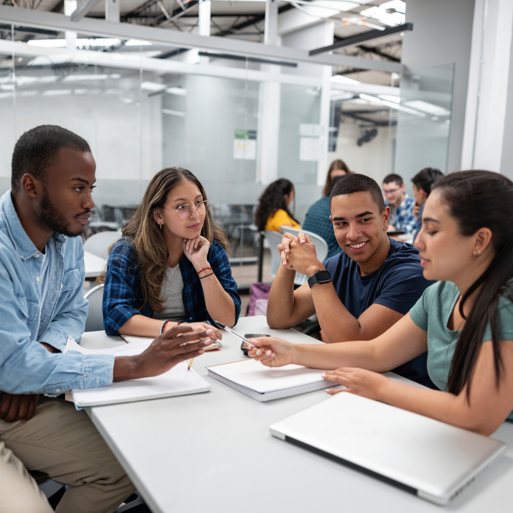Four students sit around a table in a modern classroom, discussing and taking notes together, with notebooks and pens in front of them.