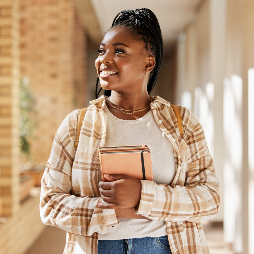 A young woman stands in a hallway holding notebooks, smiling and looking to the side. She wears a plaid shirt and has a backpack on her shoulders.