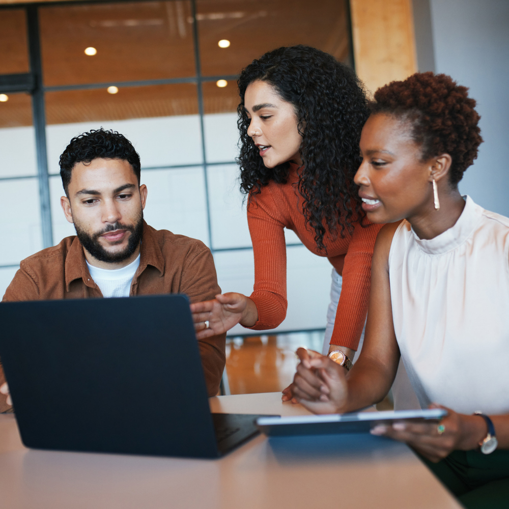 Three people collaborate at a desk, looking at a laptop screen. One person is seated typing, while the others are engaged and discussing.
