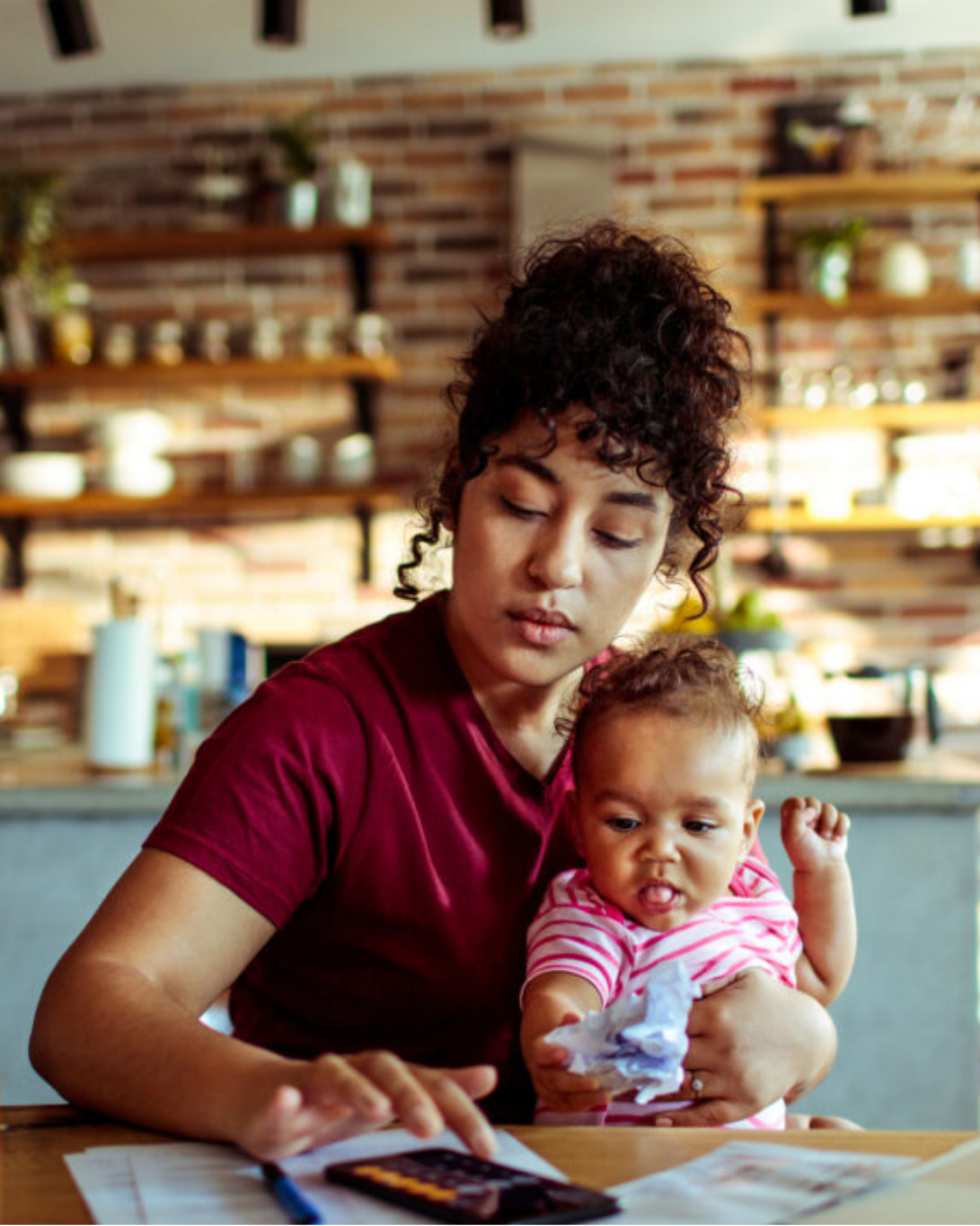 A woman holds a baby in one arm while using a calculator and reviewing papers at a kitchen table.