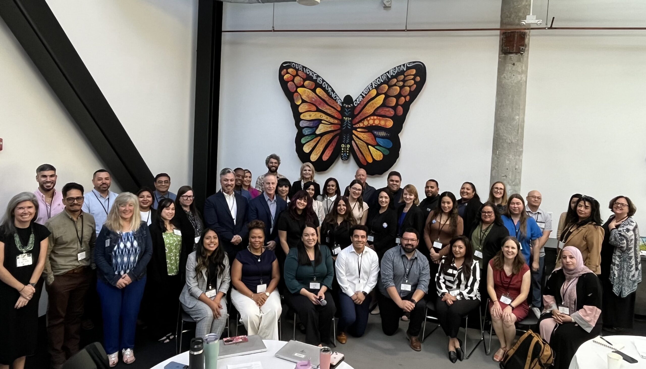 A large group of people pose and smile for a photo in front of a colorful butterfly mural in an indoor setting.