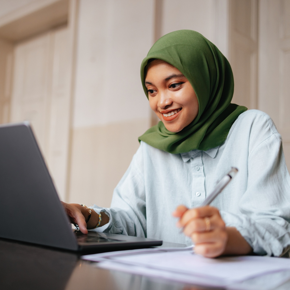 A woman wearing a green hijab works at a laptop while holding a pen and taking notes on paper at a desk.