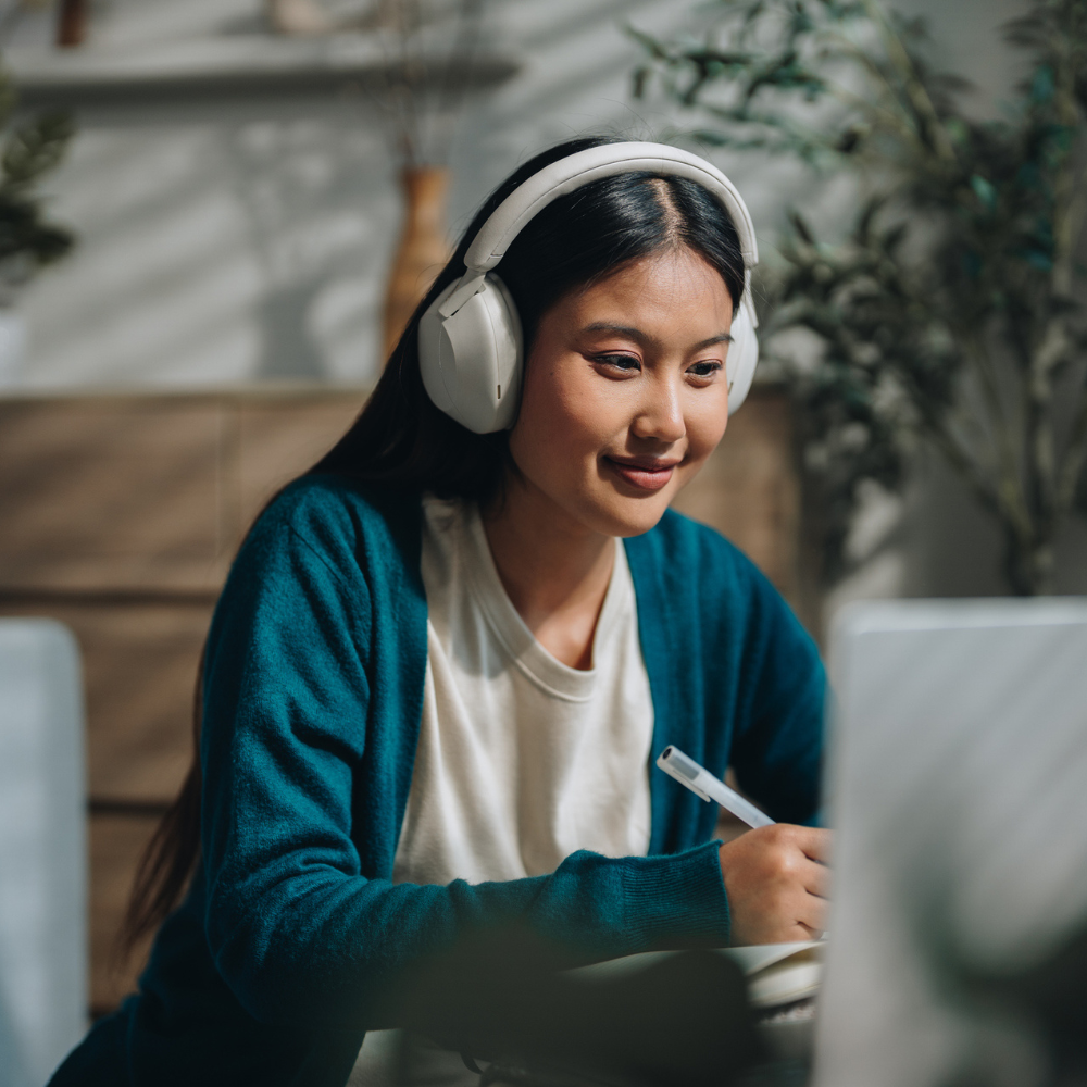 A woman wearing headphones sits at a desk, writing in a notebook while looking at a laptop screen.